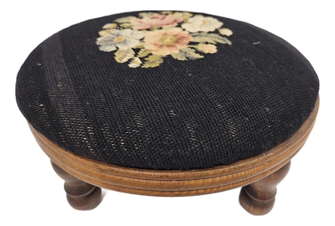 Round Needlepoint Victorian Footstool: This Victorian style round needlepoint footstool features a floral motif against black background. The base of the stool has carved lines or grooves along the outer edge. The stool is in very good vin