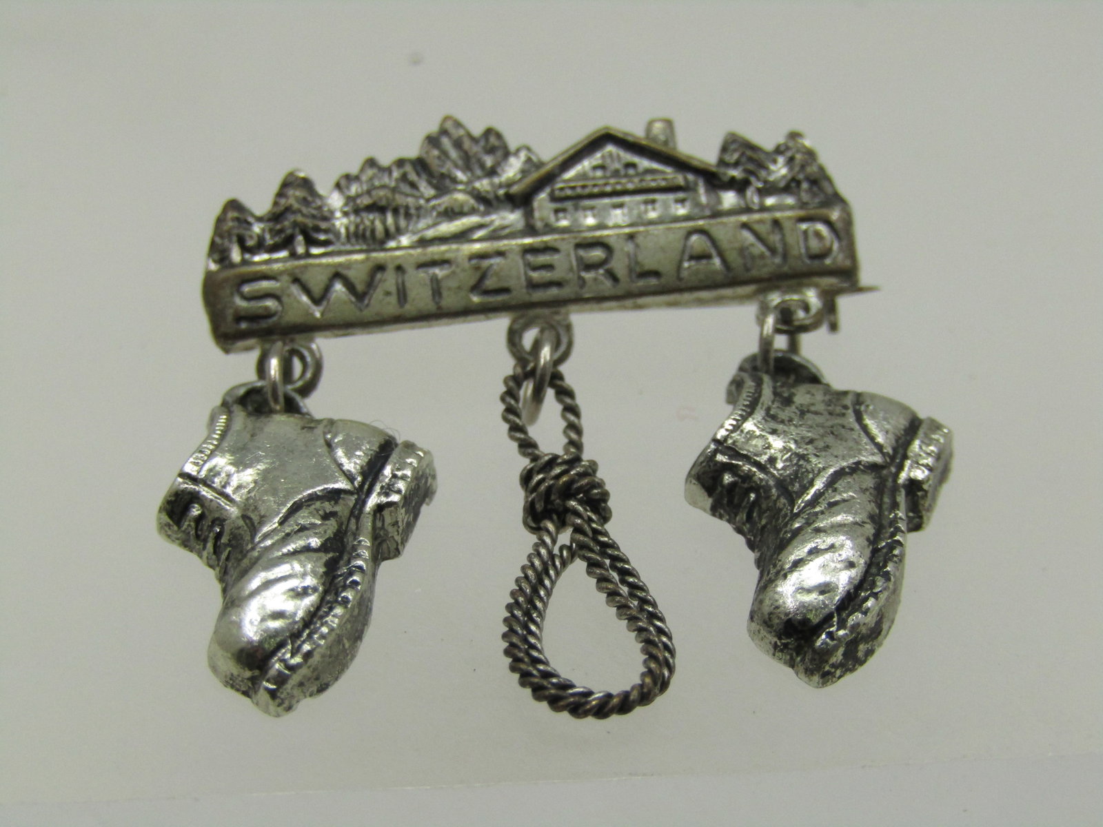 Vintage Switzerland Brooch, Village Scene with Dangles, 1.25": Vintage Swtizerland Brooch, Village Scene with Dangles, 1.25" and 1-3/8" Long. Hiker's dangles, two boots and a hope hanging from a village scene with Swizerland under that. Silver tone metal with