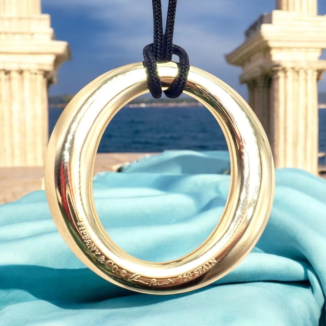 Authentic! Tiffany & Co 18k Yellow Gold Peretti 35mm Medium Sevillana Necklace: This authentic Tiffany & Co. Sevillana pendant necklace is elegantly crafted in 18k yellow gold, featuring a sleek 35mm medium-sized oval design. Suspended from a Tiffany & Co. black silk cord, it exu