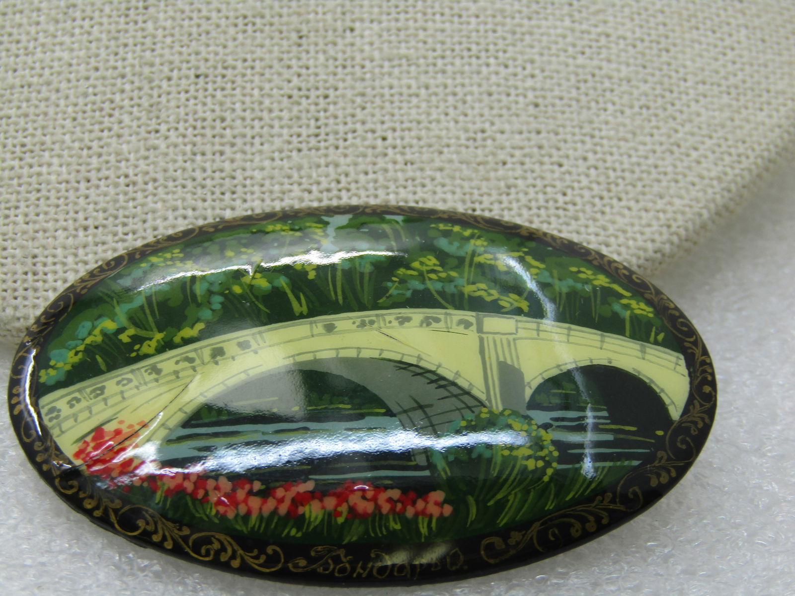 Vintage Russian Lacquered Bridge Scene Brooch, 2-1/3" Oval: Vintage Russian Lacquered Bridge Scene Brooch, 2-1/3" Oval by 1.75" tall. Green trees with pink and red flowers and a stream running under a bridge. Black background lacquered brooch with gold accents