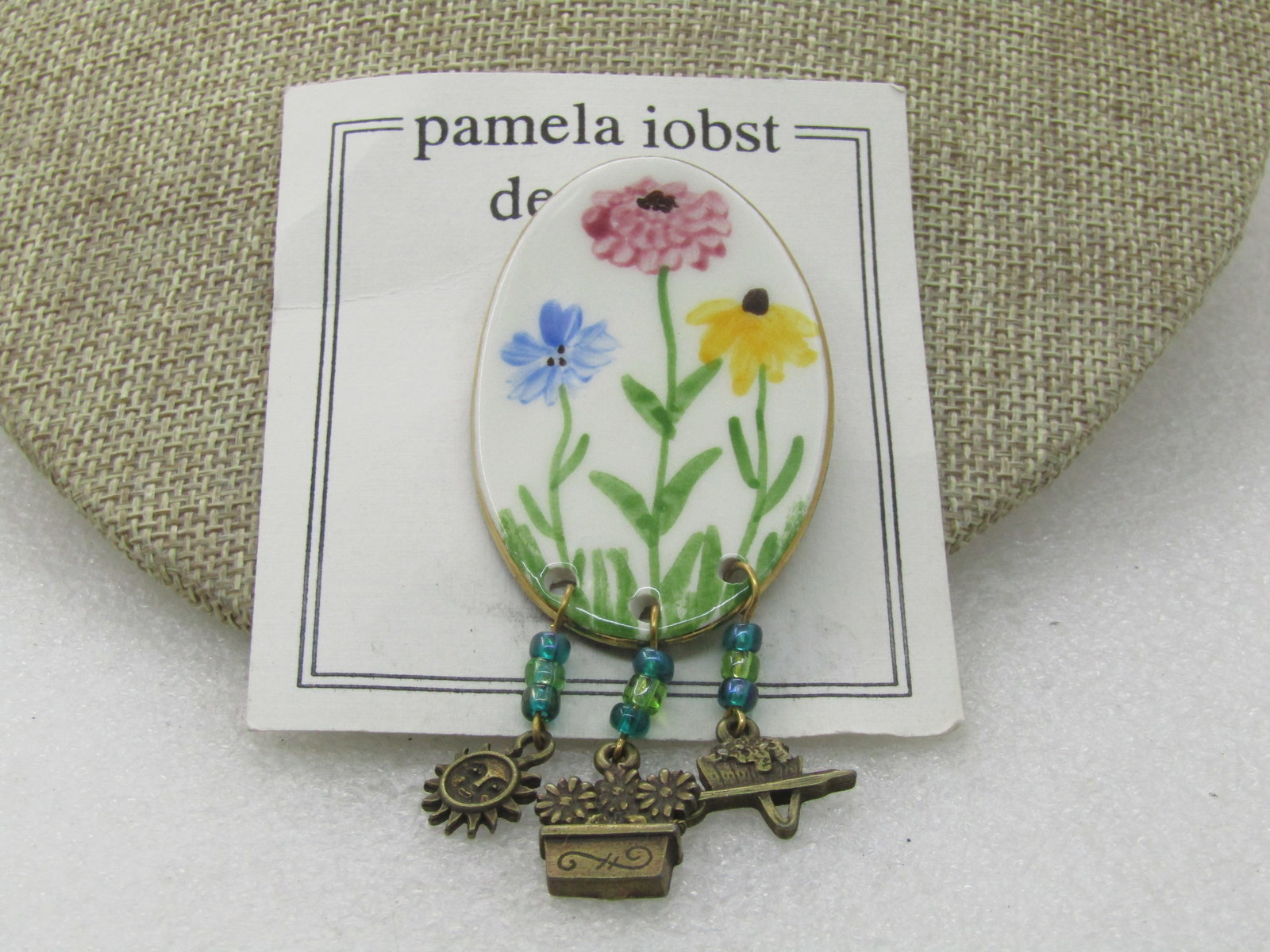 Vintage Porcelain Painted Floral Brooch with Dangles, signed Pamela Iobst Designs: Vintage Porcelain Painted Floral Brooch with Dangles, signed Pamela Iobst Designs, on original card with brass gardening dangles, flower box, sun, whellbarrow. Oval painted porcelain piece with a corn