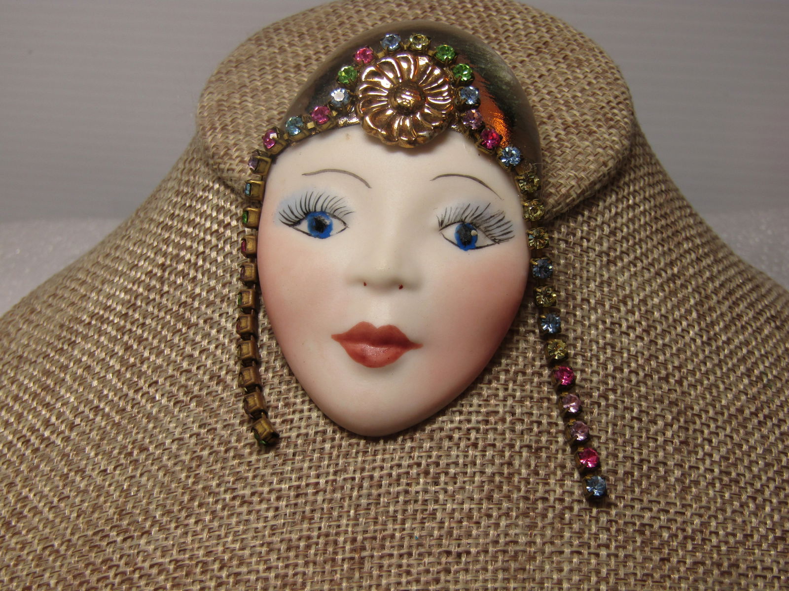 Vintage Porcelain Flapper Girl Brooch with Rhinestones, 1980's: Vintage Porcelain Flapper Girl Brooch with Rhinestones, 1980's. the face of a flapper girl with gold tone orante headpiece with blossom at the front center and a drape of pastel colored rhinestones co