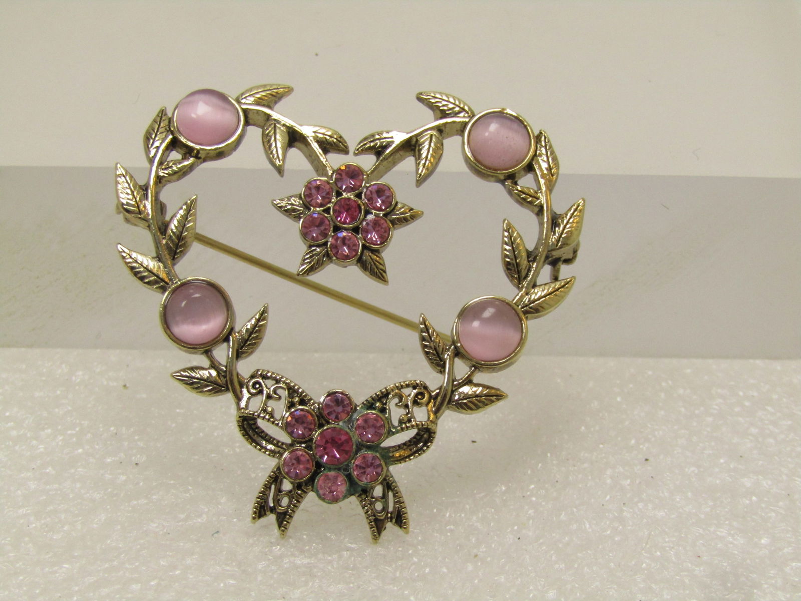 Vintage Rhinestone Heart Brooch, 1980's-1990's: Vintage Rhinestone Heart Brooch, 1980's-1990's. This is gold tone with a bow at the bottom and leaves making the frame of the heart with cateye pink/lavender round stones and light and bright pink rhi