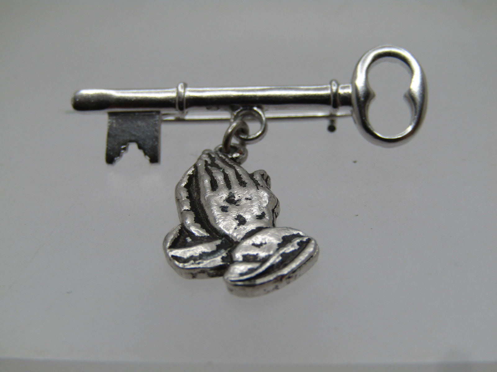 Vintage Praying Hands Key Brooch, 1960's-1970's: Vintage Praying Hands Key Brooch, 1960's-1970's. It is appx. 1.25 tall and wide. The praying hands dangle from the center of the key. The brooch is silver tone with modern c-clasp. It is not
