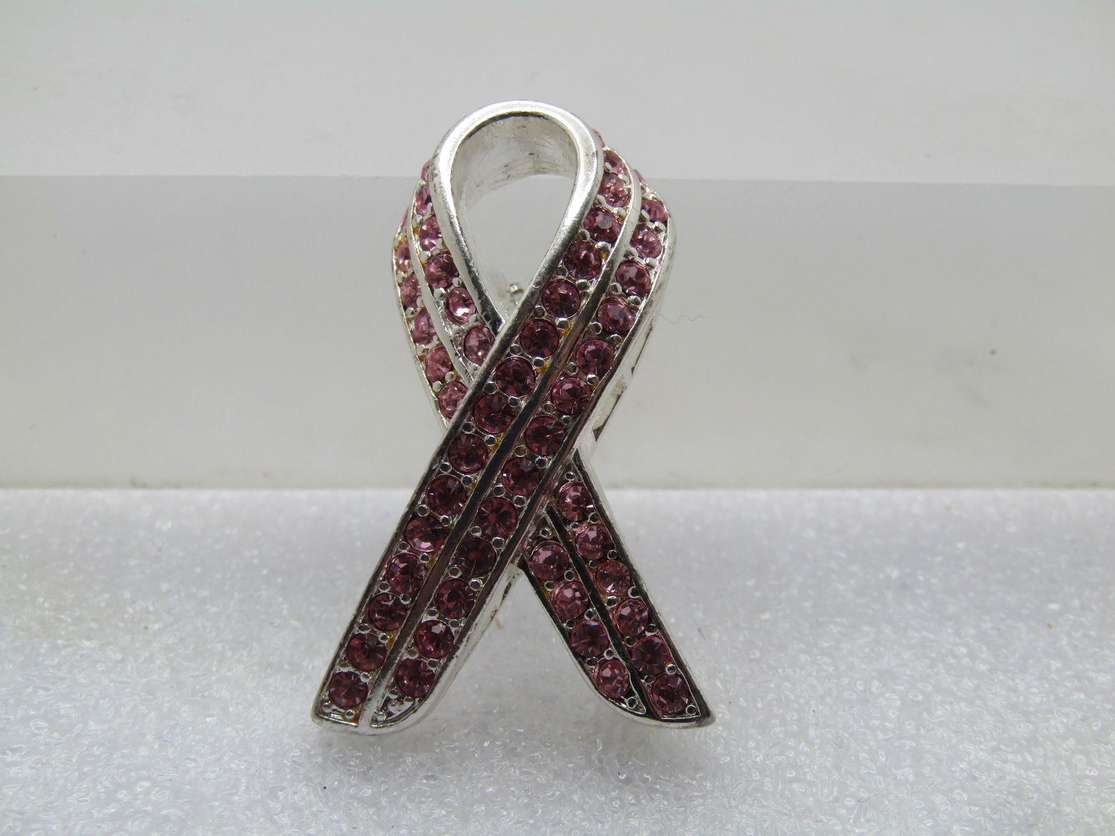 Pink Rhinestone Support Ribbon Brooch, 1990's-2000's: Pink Rhinestone Support Ribbon Brooch, 1990's-2000's. Silver tone with a double row of pink rhinestones. It has a bridge undercarriage and is not a "flat" surface brooch. It is appx 9mm tall at the