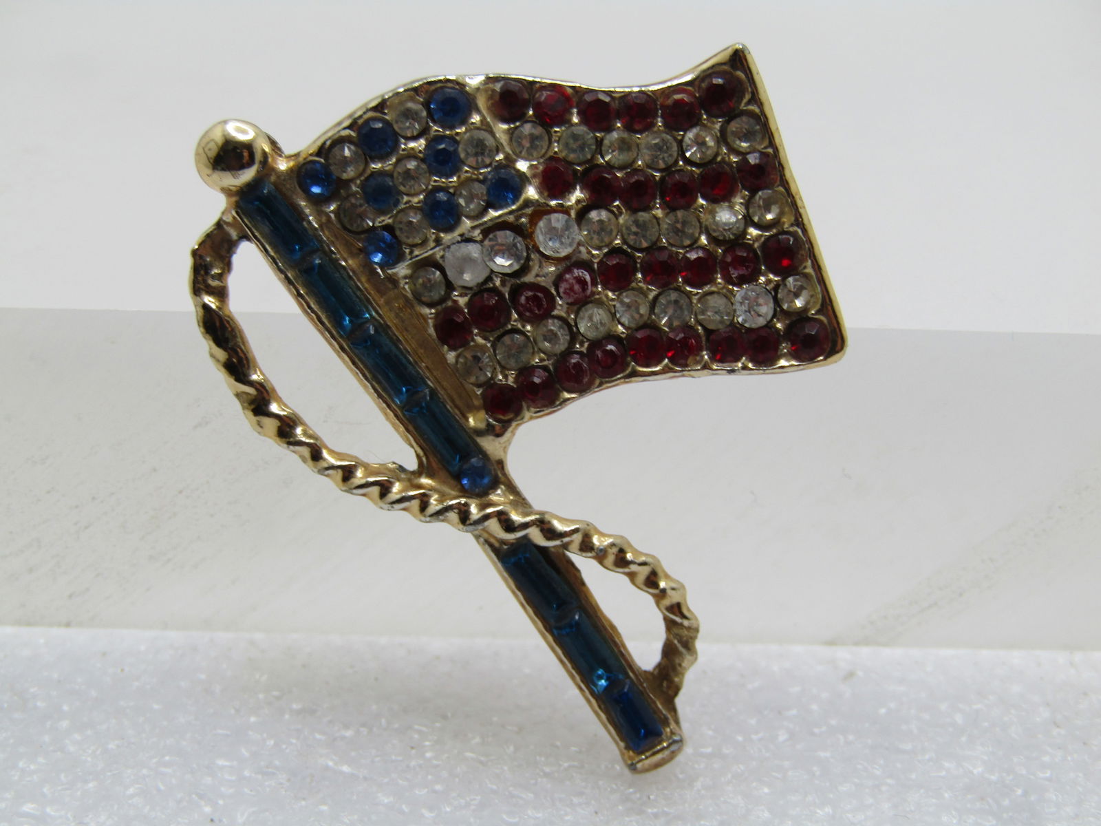 Vintage Red/Clear/Blue USA Flag Brooch 1960's-1970's: Vintage Red/Clear/Blue USA Flag Brooch 1960's-1970's. Gold tone with a wraping rope around the blue baguette pole with red, clear, and blue rhinestones set int he waving flag. It is appx. 1.75" tall