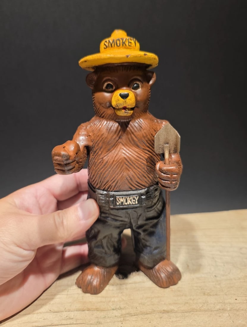 Cast Smokey The Bear Coin Bank: Title:Cast Smokey The Bear Coin BankDescription:7 7/8" High x 3 3/4" Wide Such a good reproduction cast iron bank depicting Smokey Bear, known for his role in wildfire prevention. This particular