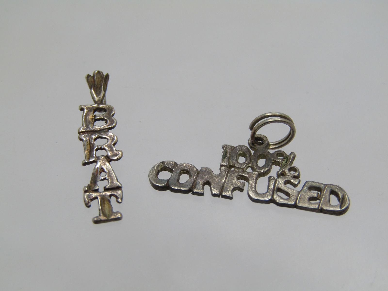 Vintage 2 Sterling Pendants 100% Confused, Brat, Signed: Vintage 2 Sterling Pendants 100% Confused, Brat, Signed NES .925 on the 100% Confused pendant - I do not see a mark on Brat; however, they both test as sterling with a combined weight of 1.36 gr. 