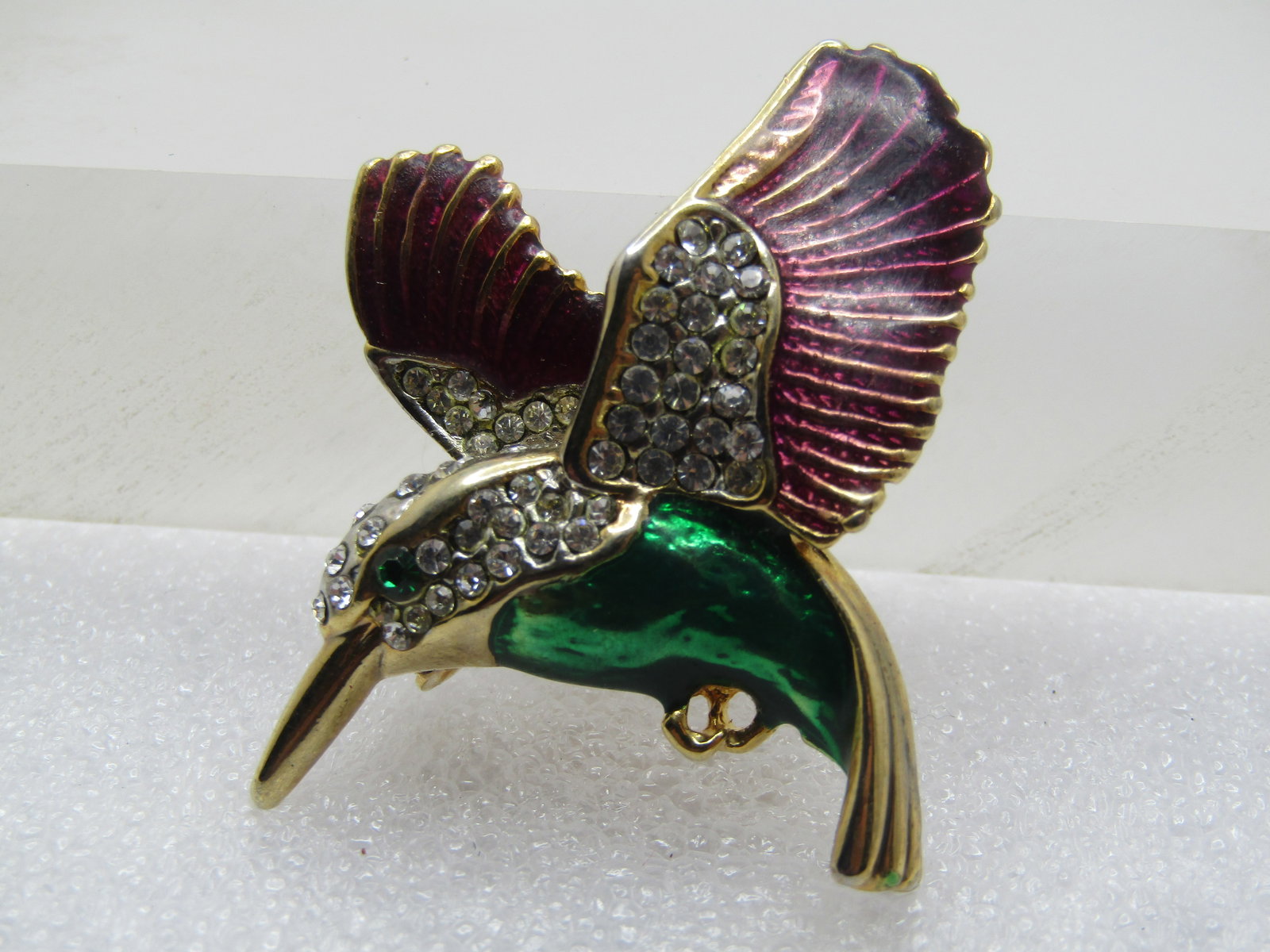 Vintage Enameled Rhinestone Hummingbird Brooch, 1980's: Vintage Enameled Rhinestone Hummingbird Brooch, 1980's, Purple wings and a green torso with green rhinestone eye and clear rhinestones in the wings and head and neck. Gold tone. It is appx. 2" by 1.75