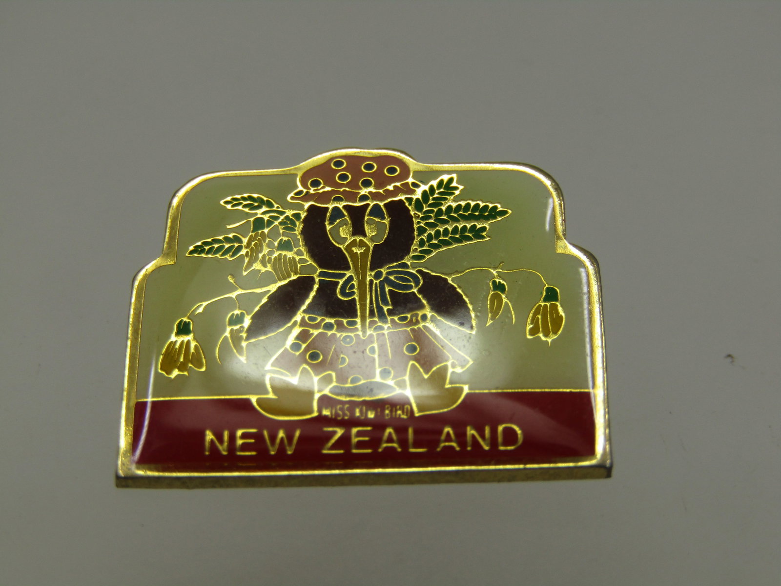 Vintage Miss Kiwi Bird Souvenier Brooch, New Zealand, Enameled,,1": Vintage Miss Kiwi Bird Souvenier Brooch, New Zealand, Enameled,,1". White background with red banner where it state New Zealand in gold lettering. A cute girl kiwi with leaves behind her and