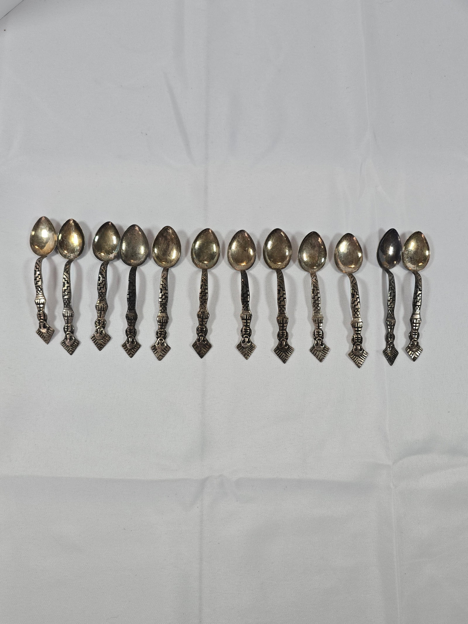 Set of Twelve Vintage Silver-Toned Decorative Spoons: A charming set of twelve vintage silver-toned decorative spoons, likely a base metal with silver plating. Each spoon features an ornate handle with geometric and figural design elements, culminating