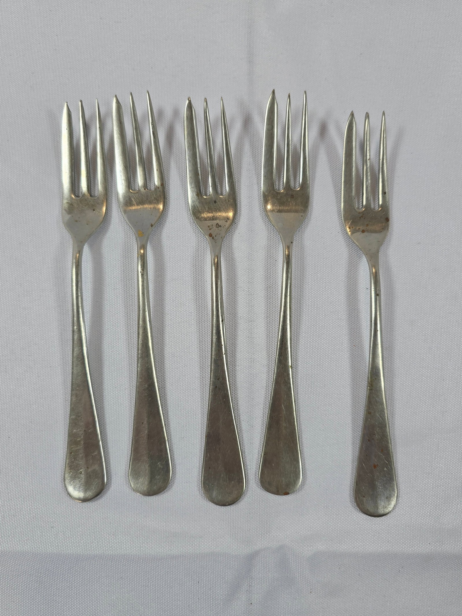 Set of Five Sterling Silver or Silver-Plated Forks: A set of five elegant forks, likely crafted from sterling silver or silver-plated metal, featuring a classic, unadorned design typical of late 19th or early 20th-century flatware. The forks exhibit a