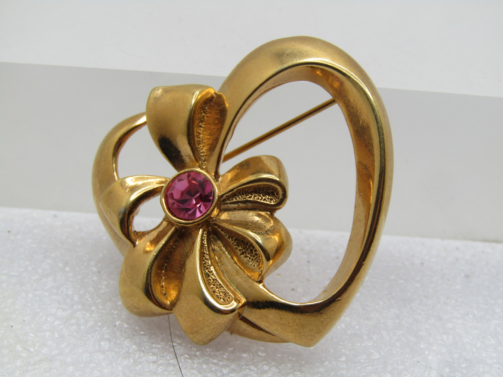 Vintage Heart & Bow Brooch with Pink Rhinestone, Avon, 1960's-1970's: Vintage Heart & Bow Brooch with Pink Rhinestone, Avon, 1960's-1970's. Gold tone and appx. 1.5" tall and wide - open heart with smooth and shiny finish and an angle frame that rises at the center. Ther