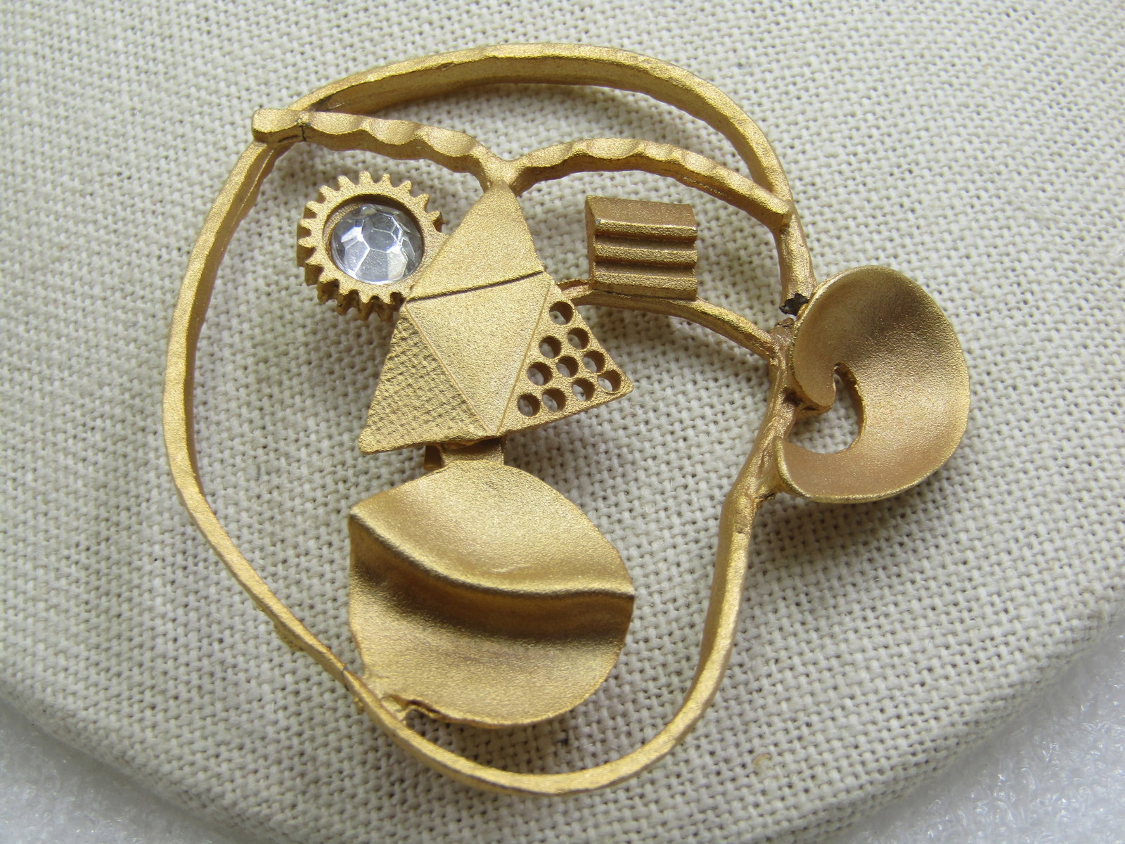 Vintage Modern Steam Punk Face Brooch, 2.75" by 2.5": Vintage Modern Steam Punk Face Brooch, 2.75" by 2.5", appx. 1980's. A single rhinestone eye with gear frame and geometric nose and eye and bend lips and a single curved ear. Satin finished gold tone