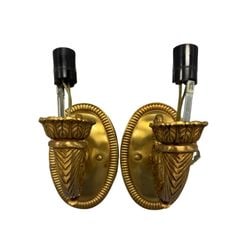 Pair of Gilt Bronze Empire Style Sconces: A pair of ornate gilt bronze sconces in the Empire style, featuring Neoclassical motifs. Each sconce is comprised of an oval backplate with a reeded border, supporting a torch-form arm adorned with ac