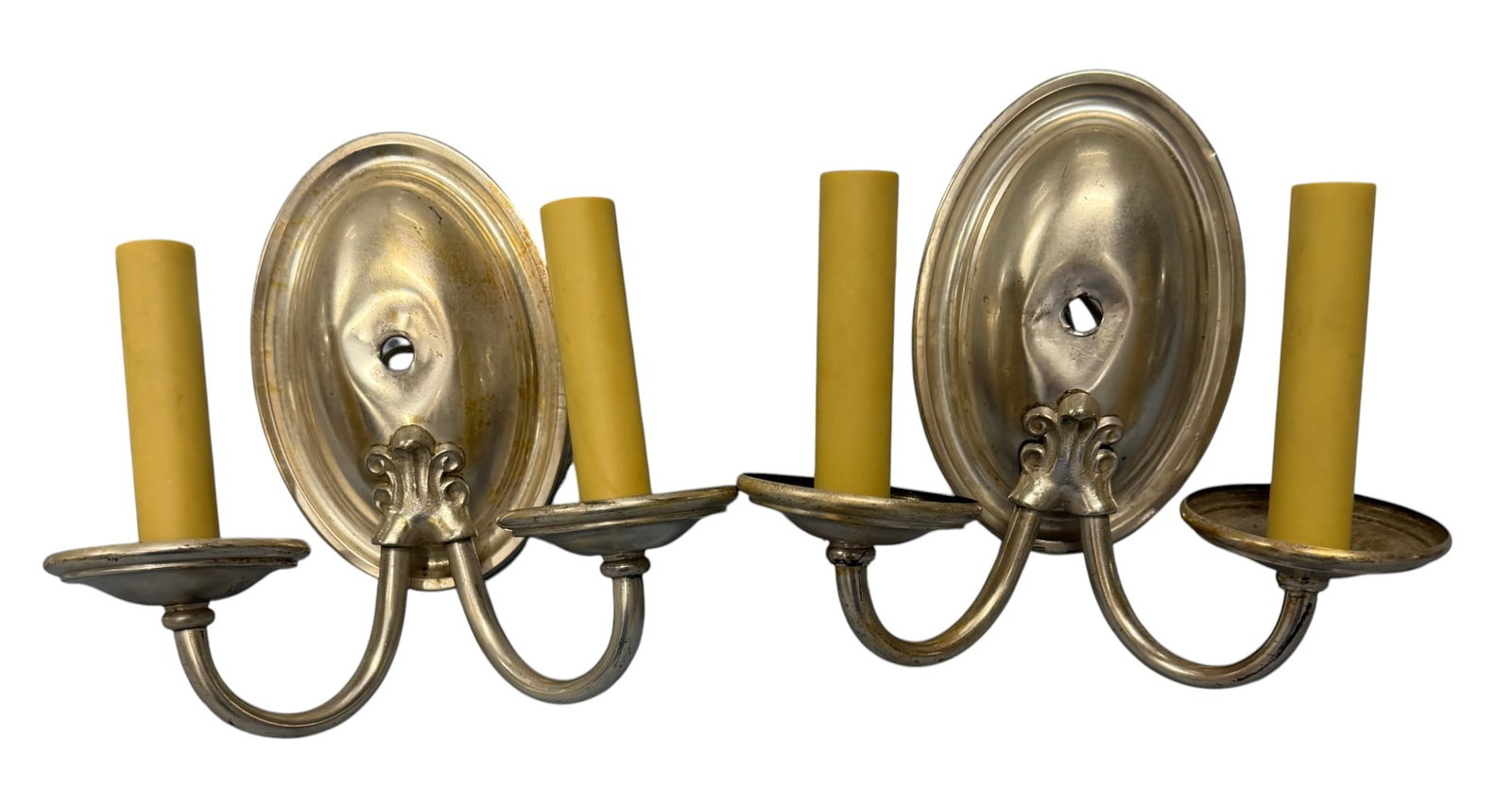 Pair of Neoclassical Style Silver Plated Metal Wall Sconces: A pair of Neoclassical style silvered metal wall sconces, likely mid-20th century. Each sconce features an oval backplate decorated with a scroll and acanthus leaf motif at the base, from which two