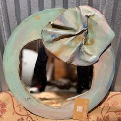 Hand-Painted Abstract Ceramic Wall Mirror with Ruffled Accent: This unique ceramic wall mirror features a circular, hand-painted frame in a pastel palette of muted green, pink, and blue, suggesting an abstract or contemporary art style. A prominent, ruffled