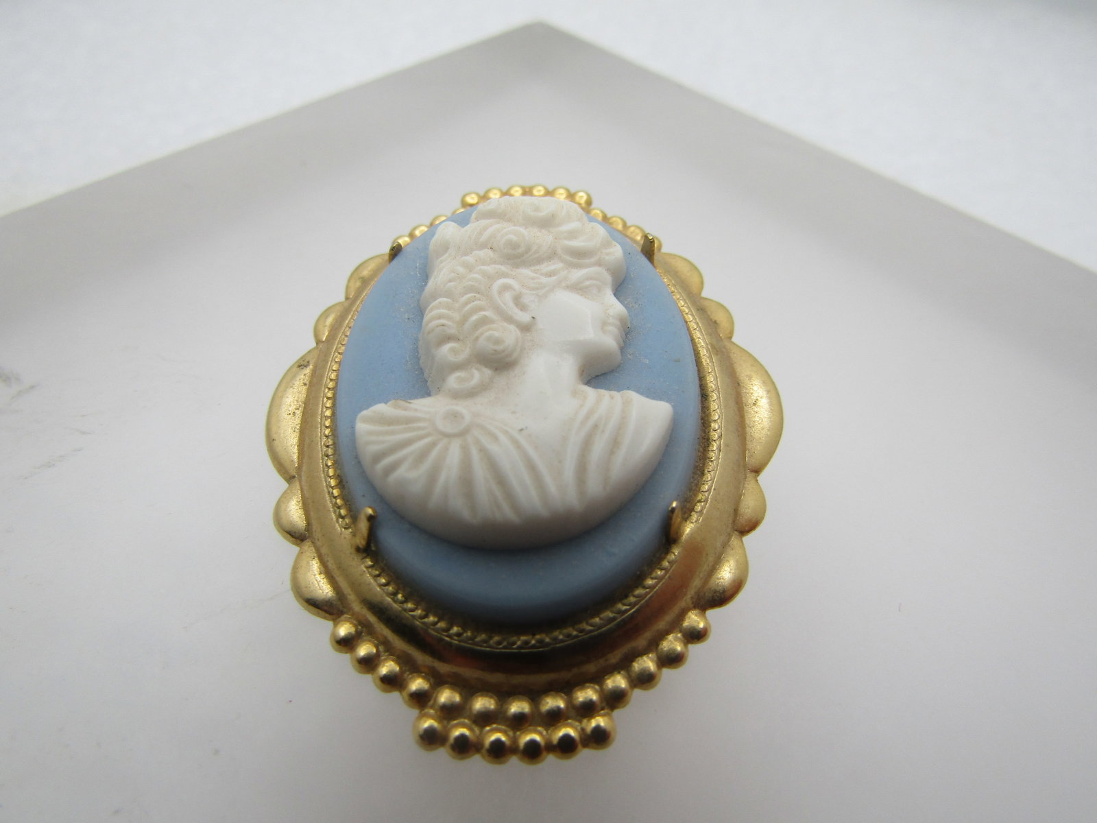 Vintage Faux Jasperware Cameo Brooch, 1960's-1970's: Vintage Faux Jasperware Cameo Brooch, 1960's-1970's. Blue and white oval cameo with the jasperware colored background and a pressed white cameo on top. Tiered gold tone frame. Locking clasp. Gold tone