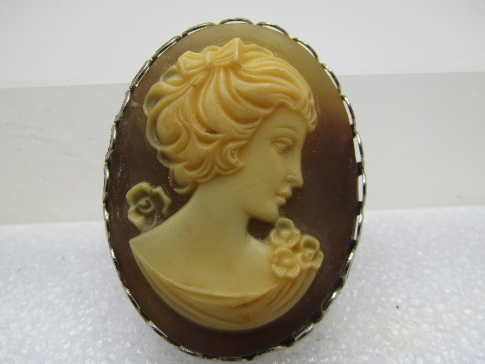 Vintage Cameo Brooch, 1960's-1970's, 1.75" by 1.25": Vintage Cameo Brooch, 1960's-1970's, 1.75" by 1.25". Molded cameo with a brownish background and offwhite cameo with floral accents, curlved hair and bow. Silver tone. , locking clasp. Very good and c