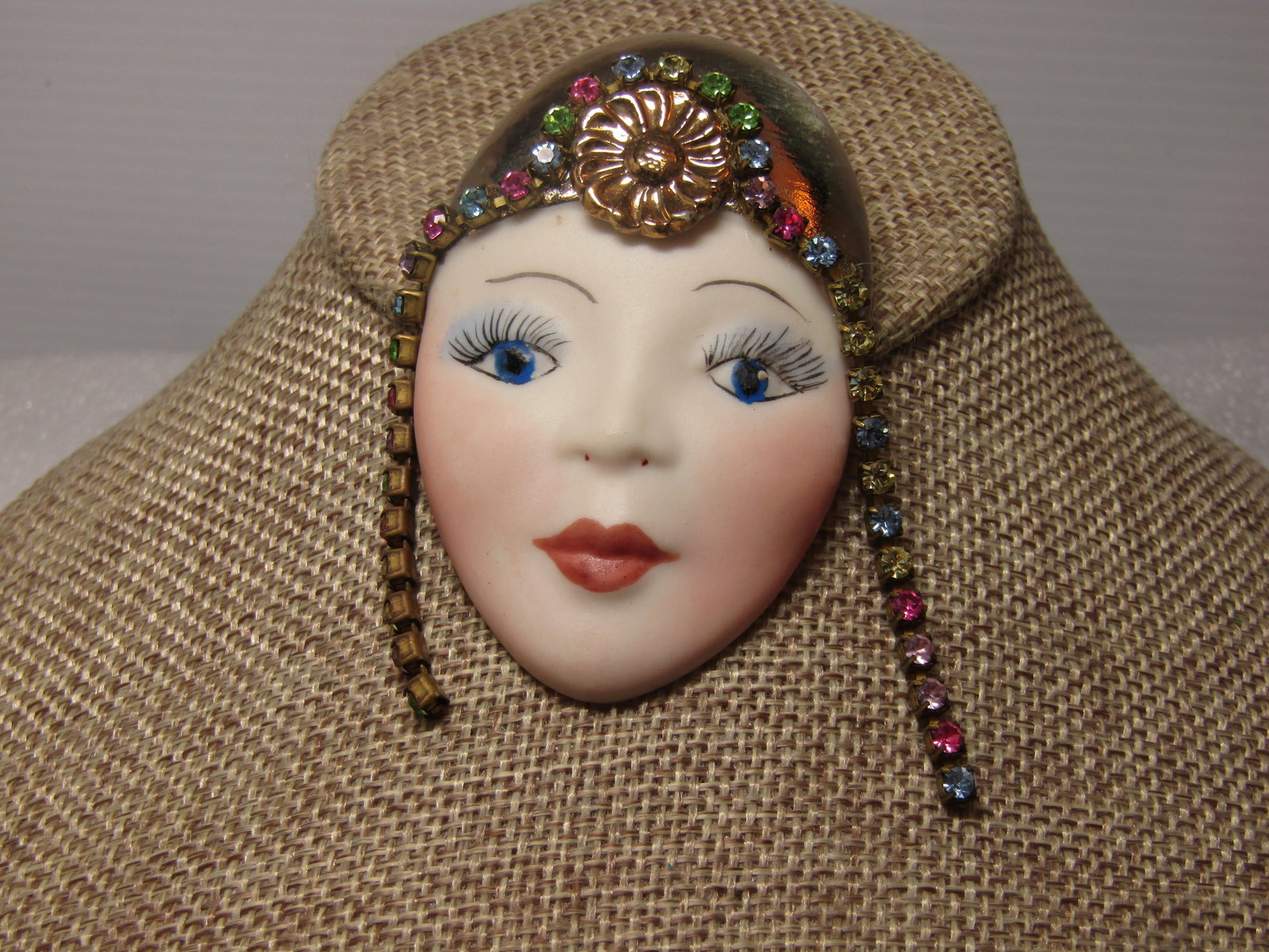 Vintage Porcelain Flapper Girl Brooch with Rhinestones, 1980's: Vintage Porcelain Flapper Girl Brooch with Rhinestones, 1980's. the face of a flapper girl with gold tone orante headpiece with blossom at the front center and a drape of pastel colored rhinestones