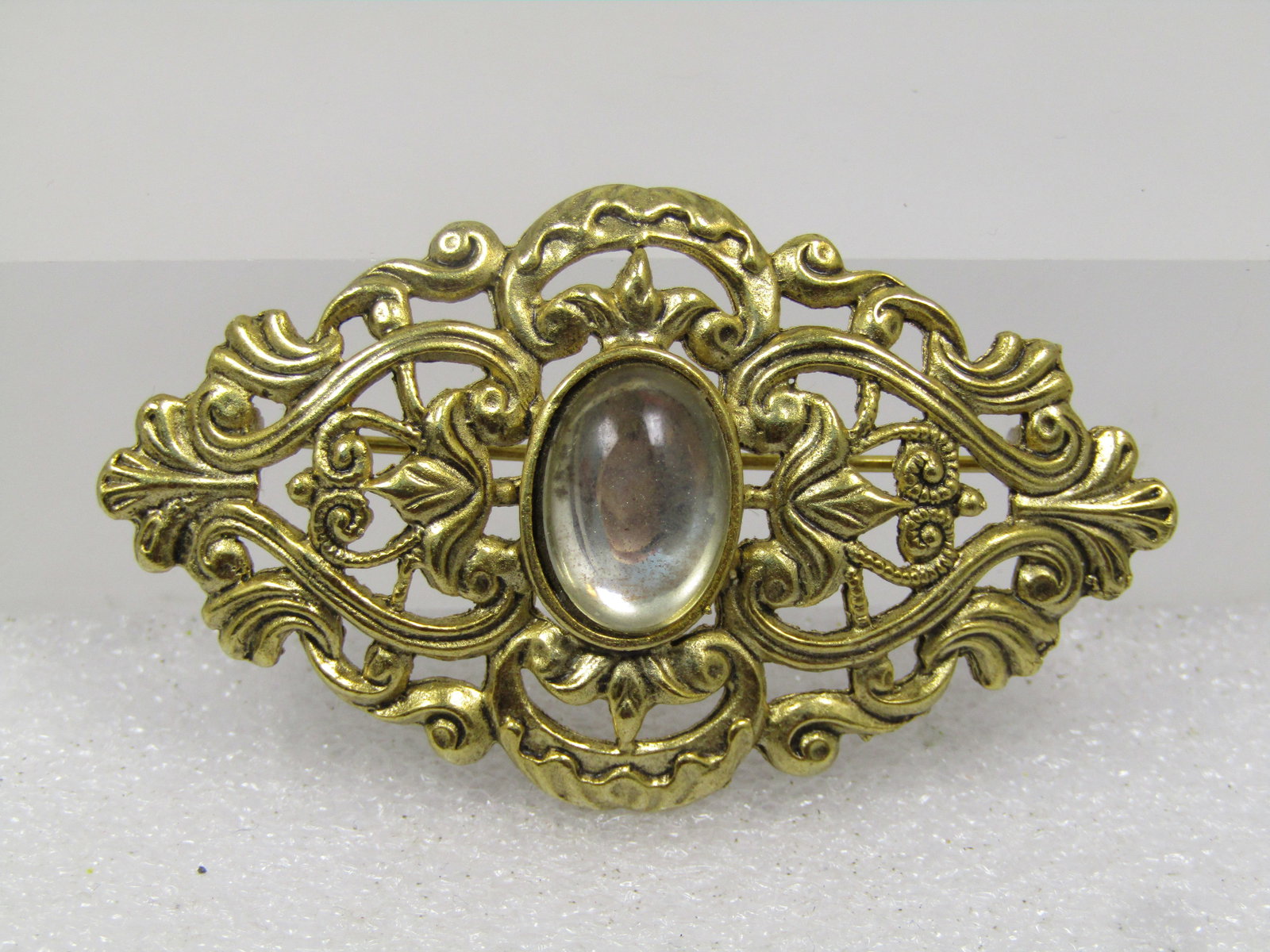 Vintage Gold tone Victorian Themed Brooch, Clear Glass Stone, 1980's: Vintage Gold tone Victorian Themed Brooch, Clear Glass Stone, 1980's. The brooch is gold tone with scrolled design with lower and higher areas that flow from side-to-side. The center is a clear glass 