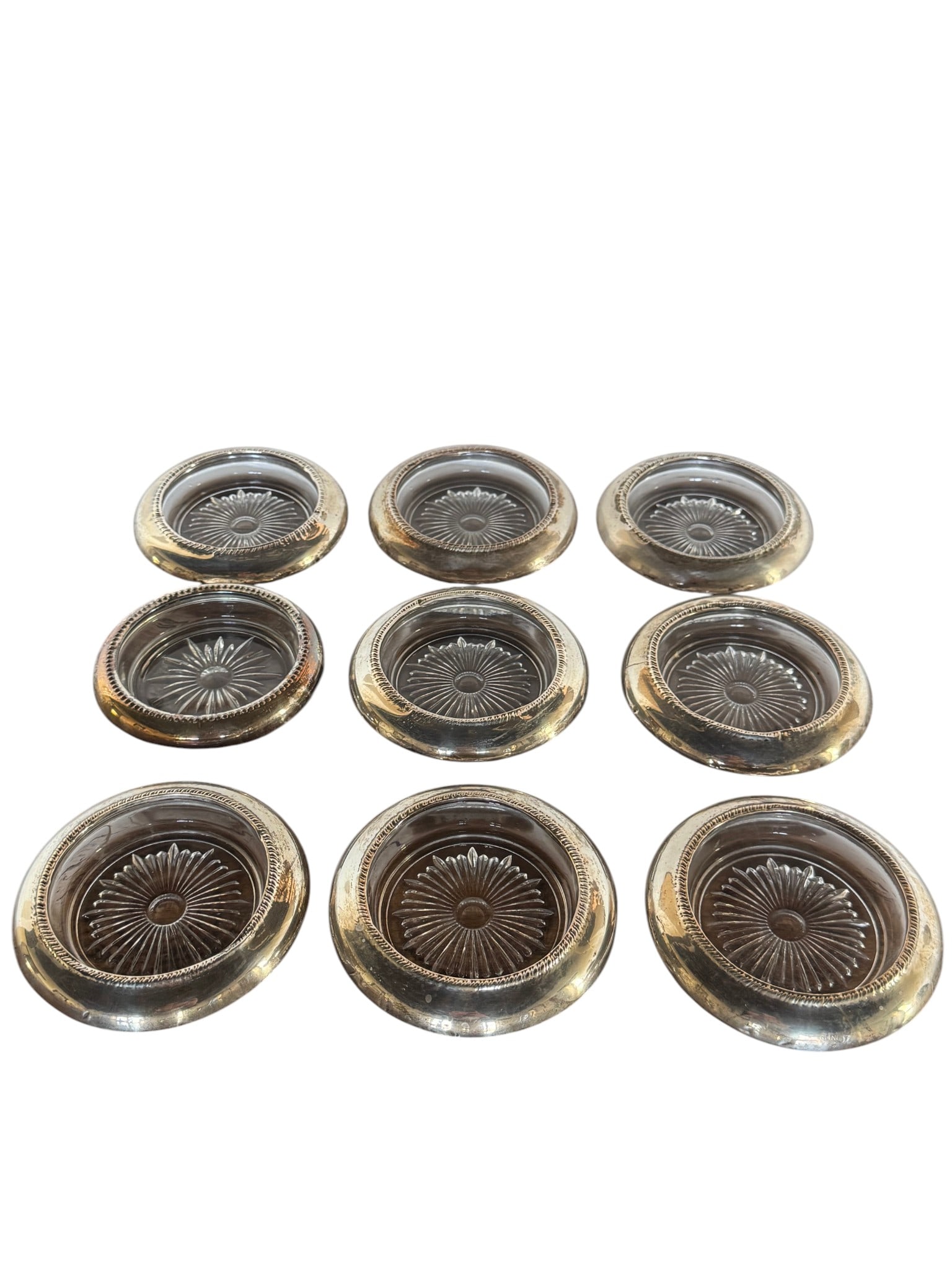Set of Nine Silver Plate and Glass Coasters: A set of nine elegant coasters, each featuring a silver-plated rim with a reeded edge surrounding a clear glass insert. The glass inserts are circular with a starburst pattern molded into the center,