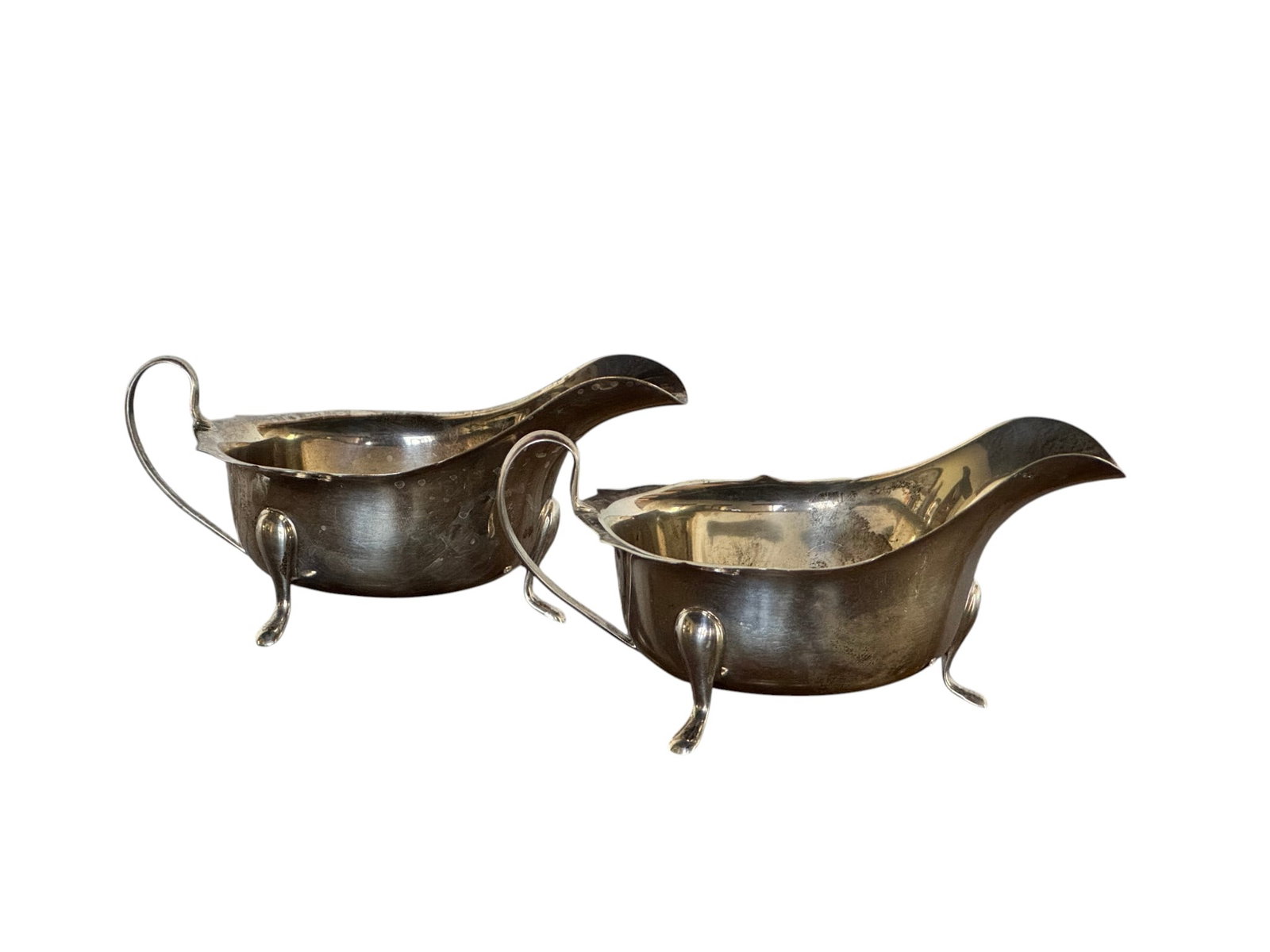 Pair of Sterling Silver Gravy Boats: A pair of sterling silver gravy boats. Each boat features a classic oval body with a graceful pour spout, an elegant reeded handle, and stands on three delicate hoofed feet. The underside of each