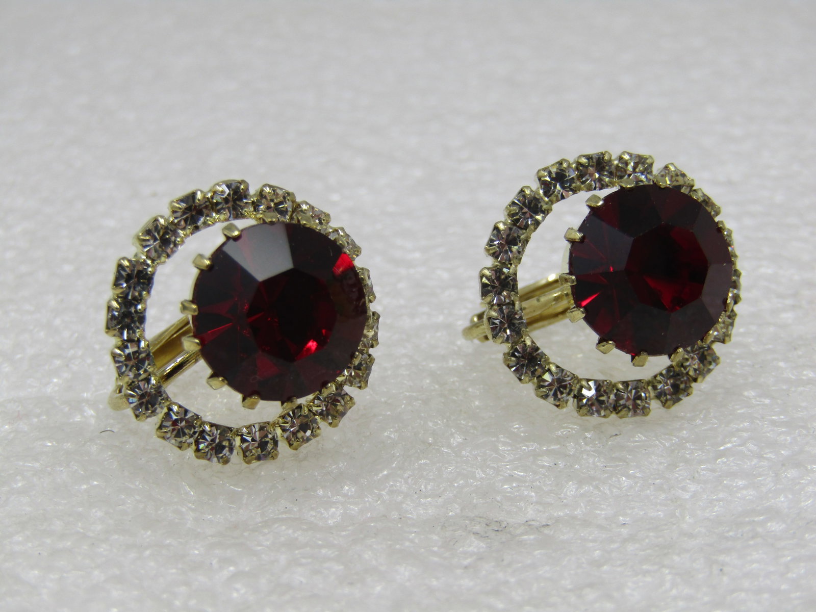 Vintage Red Clear Rhinestone Earrings, Hinged Screwback: Vintage Red Clear Rhinestone Earrings, Hinged Screwback. Appx. 11mm round red rhinestone with an offset halo around the stone with open space. Top of the earring is appx. 18mm round. Gold tone with a 