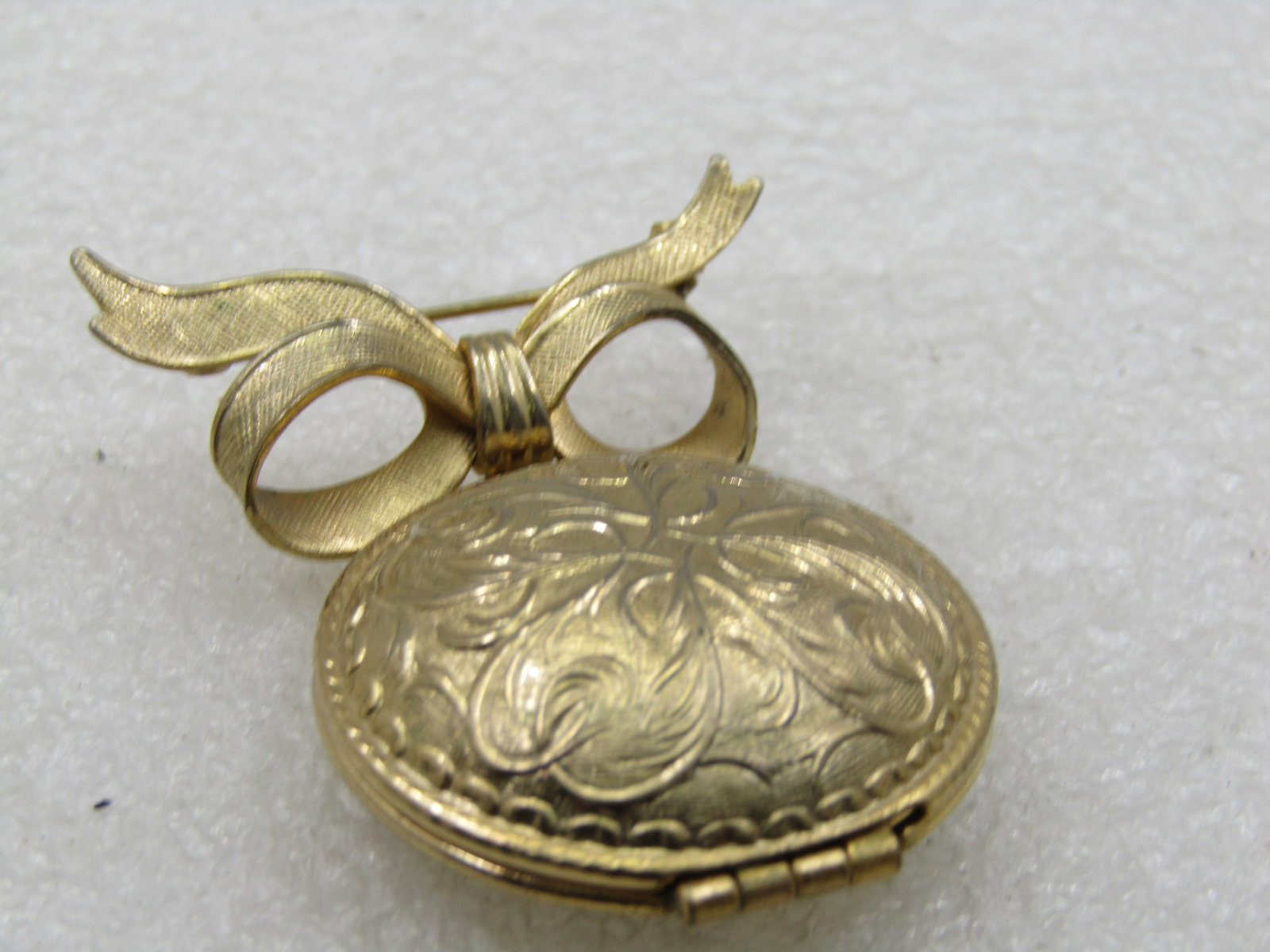 Vintage Avon Bow & Perfume Locket Brooch, 1970's-1980's: Vintage Avon Bow & Perfume Locket Brooch, 1970's-1980's. Gold tone round domed locket with raised scrolled design and a textgured bow. The label on the back is Avon Prod Inc. Unforgettable. New York,