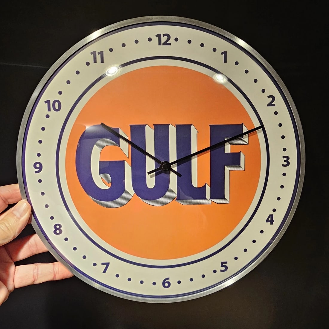 12" Round Gulf Oil Wall Clock Sign: 12" Wonderful reproduction vintage-style "Gulf" branded wall clock, featuring a circular orange face with the blue "GULF" logo prominently displayed. It has a black outer casing, standard clock