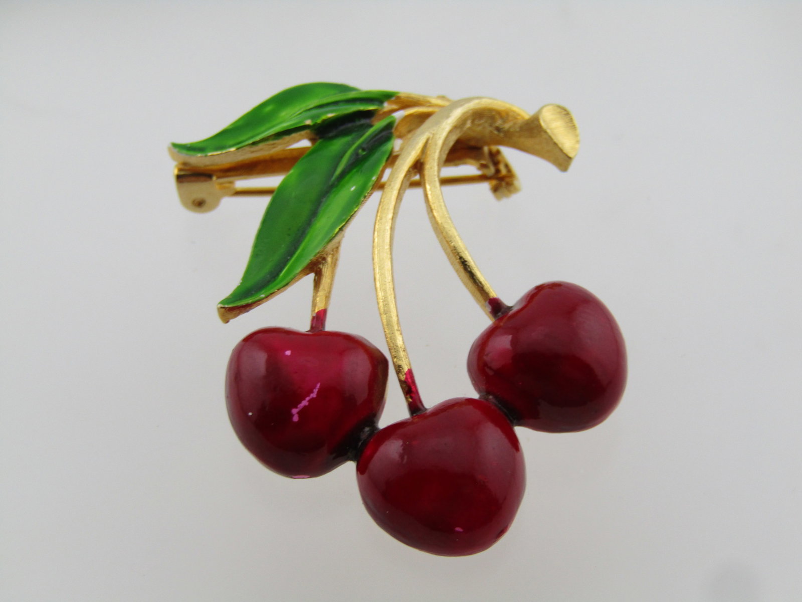 Vintage Enameled Cherry Stem Brooch, 1960's-1970's: Vintage Enameled Cherry Stem Brooch, 1960's-1970's. Gold tone with two green enameled leaves and three cherries. The cherries curve on their stems. They look over the stem on top with the leaves offse