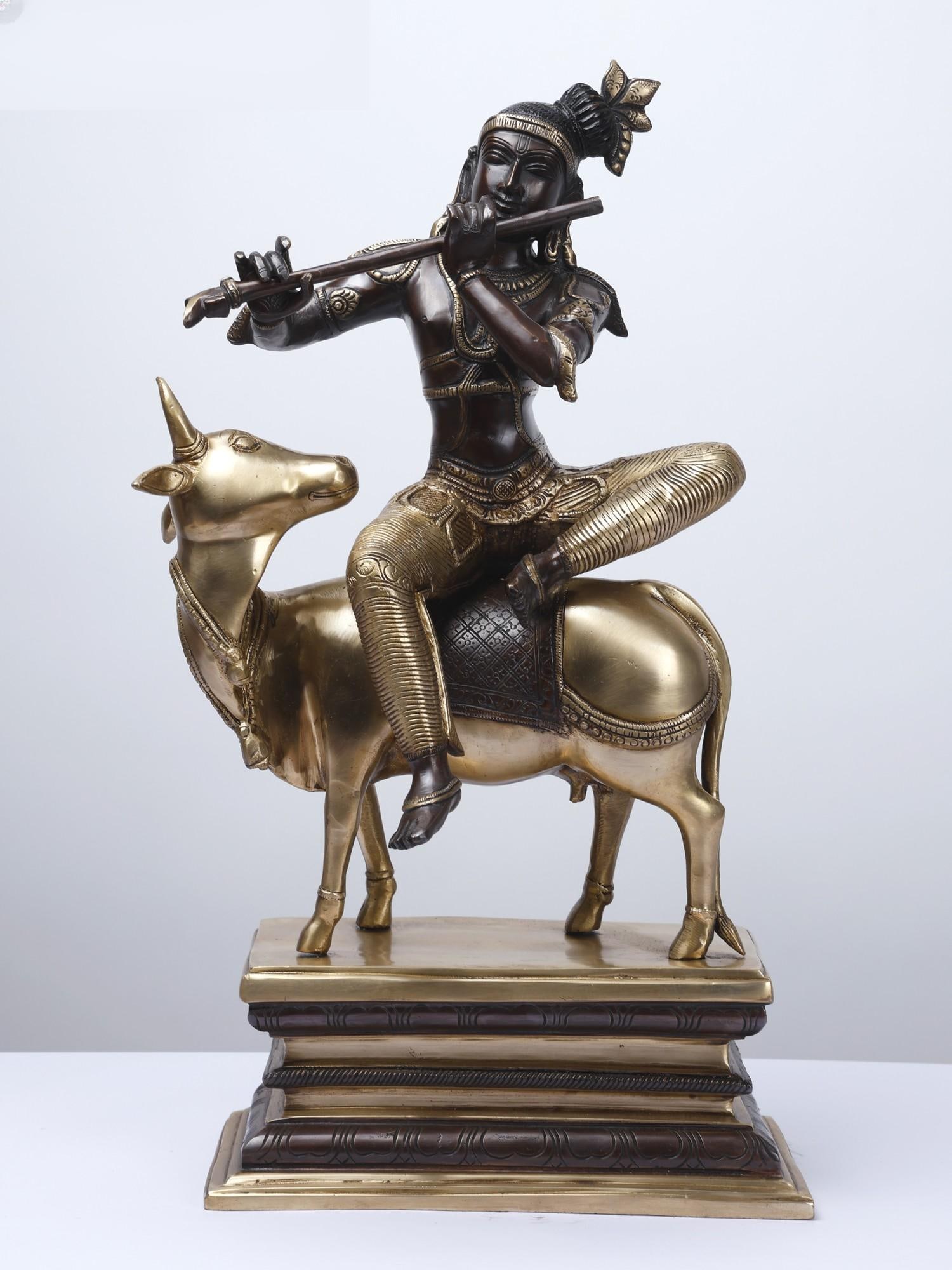 20" Fluting Krishna Seated on Cow /Gopala Krishna Brass Statue: Description: On the banks of Yamuna, Krishna plays his flute, mesmerizing his cattle folk, residents of Vrindavana, and the entire Universe. An ode to Krishna’s bonds with all living beings of the w
