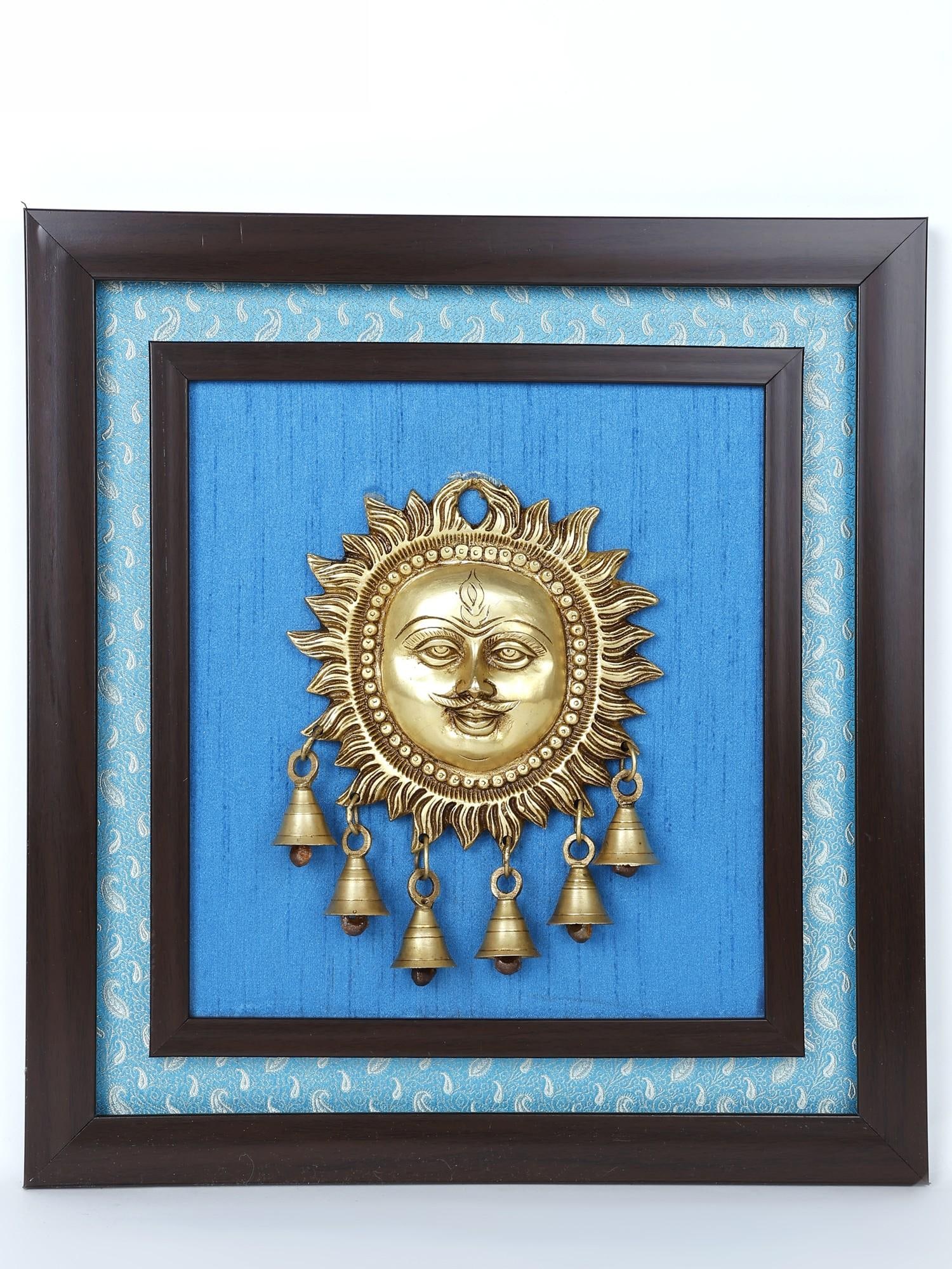 15" Lord Surya with Dangling Bells in Brass /Wood Framed Brass Sculpture /Wall Hanging Decor: Description: Surya- the resplendent ancient deity is presented in this brass and wood wall hanging in a traditional iconography, against a textured cloth background that has a modern appeal. The Hindu