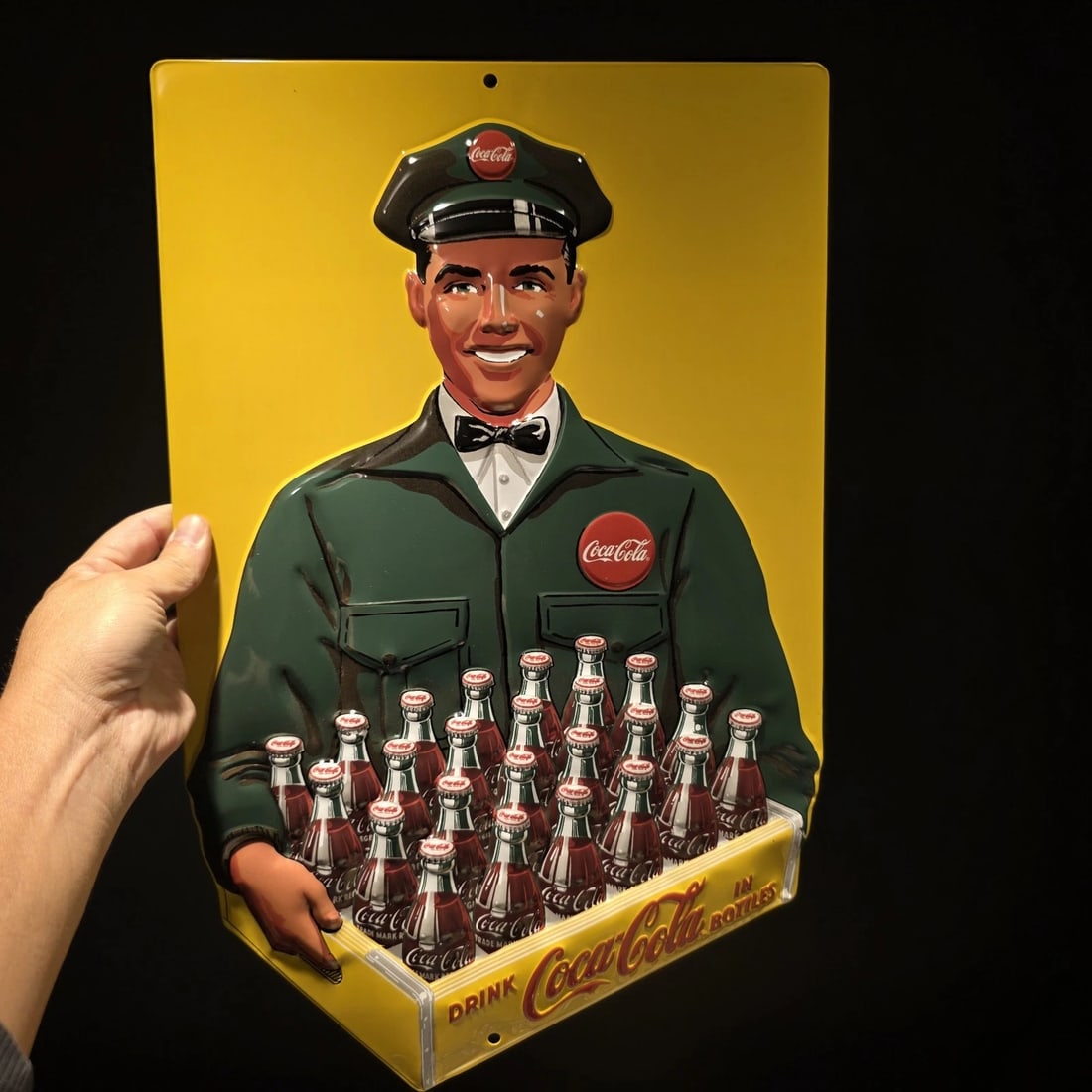 20" Coca-Cola Delivery Man Sign Embossed Coke Advertising: 12 1/2" x 20" Embossed Coca-Cola advertising sign by Ande Rooney, featuring a uniformed delivery man holding a case of glass Coke bottles. Ande Rooney signs are known for their layered embossing, roll