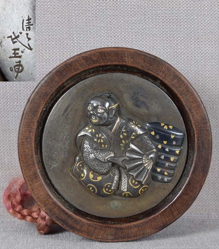 19c netsuke ACTOR with boxes by MINGYOKU: 19th century kagamibuta (“mirror lid”) netsuke with multimetal top and wooden bowl. The top is inlaid with an actor wearing a mask and holding an open fan in his hand seated next to an