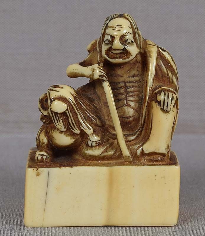 18c netsuke SENNIN IZENSHUN WITH DOG: 18th century netsuke of an emaciated sennin seated on a base wearing a tattered robe and a leaf cape and holding a staff with a bundle of his possessions tied to its top, and with a dog affectionately