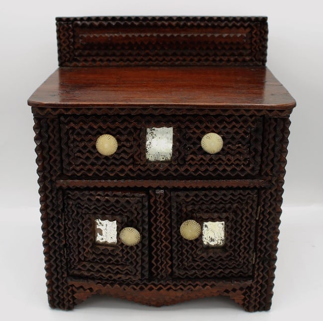 A very well made tramp art miniature dresser: This is a tramp art miniature dresser. It has a drawer over two doors, decorated with flatwork, with three small mirrors set in. The surface is a dark, glossy lacquer or varnish. Inside are color
