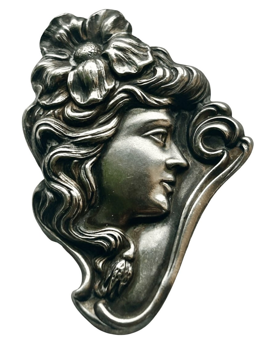 Art Nouveau Style Silvered Metal Brooch with Female Profile: An elegantly sculpted Art Nouveau style brooch crafted from silvered metal, featuring the profile of a young woman adorned with a floral wreath or elaborate coiffure. Her flowing hair and the organic