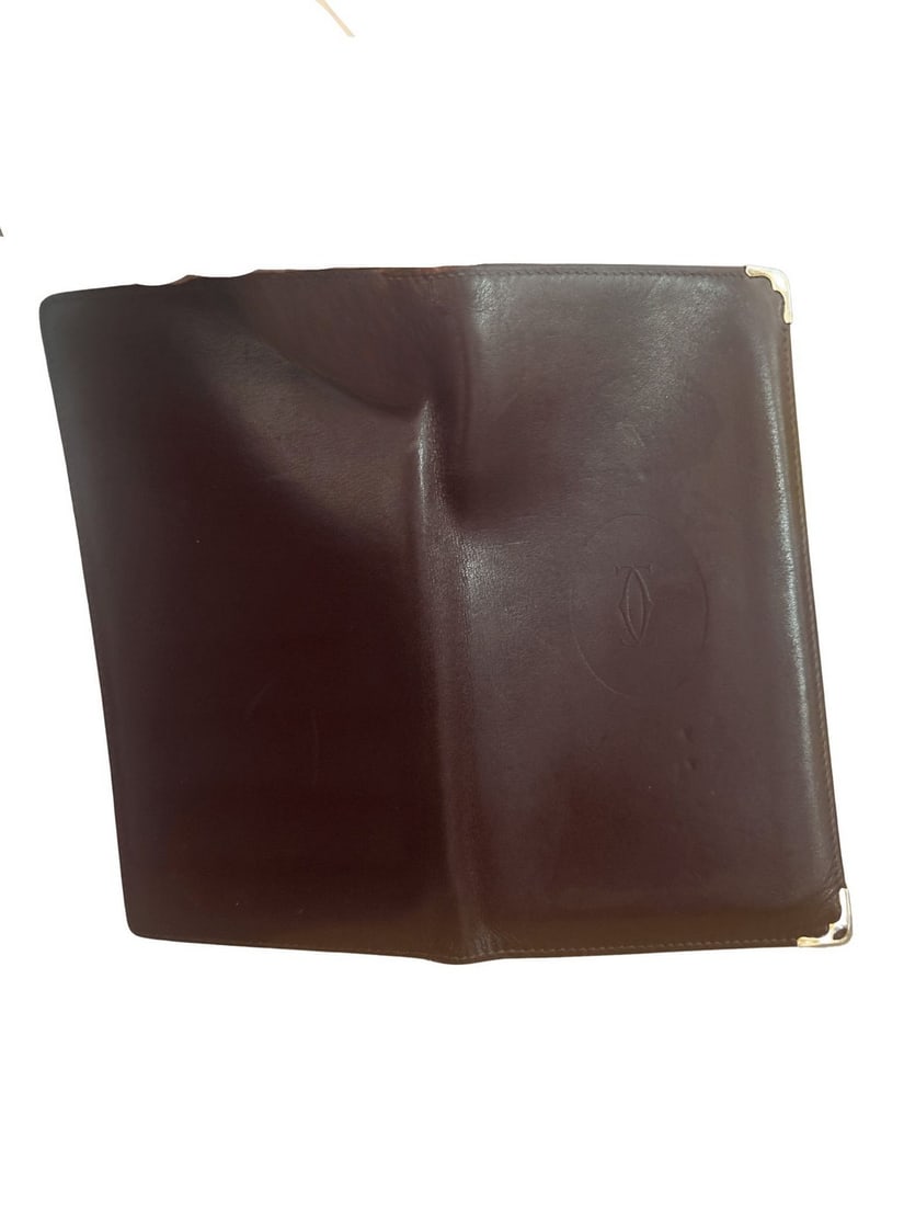 Cartier Paris Bi-Fold Wallet in Burgundy Leather: A sophisticated Cartier Paris bi-fold wallet crafted from rich burgundy leather, featuring gold-toned metal corner accents and the embossed Cartier logo on the exterior and interior. The interior is