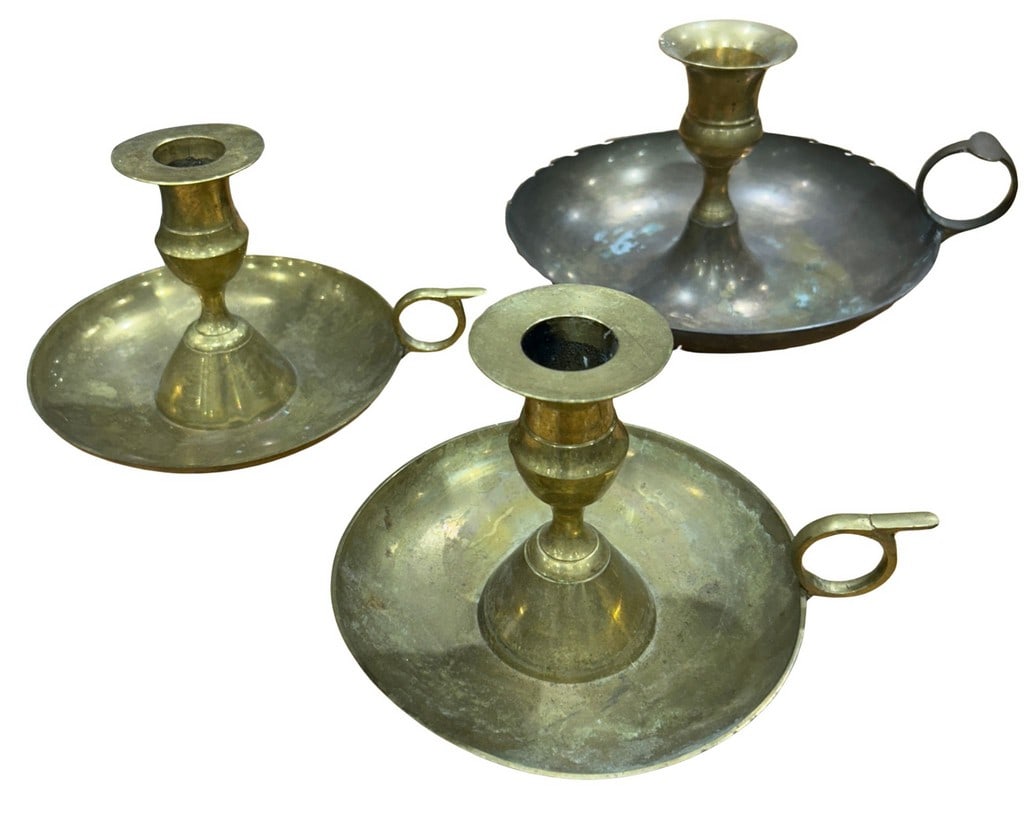 Set of Three Vintage Brass Handled Chamber Candlesticks: This lot features a set of three vintage brass chamber candlesticks, two in polished brass and one with a silvered finish. Each candlestick is designed with a broad drip pan to catch wax and a finger 