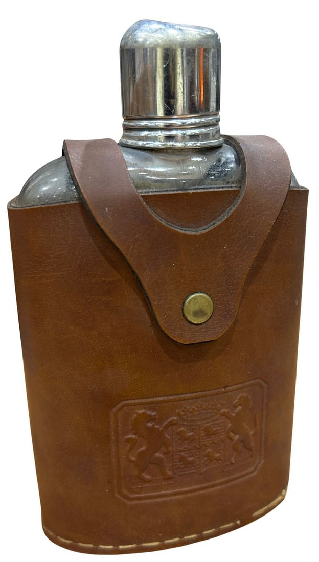 Glass Flask in Embossed Leather Case: A clear glass flask housed in a fitted brown leather carrying case. The leather case features an embossed heraldic crest design on the front, snap closure, and visible stitching along the edges. The f