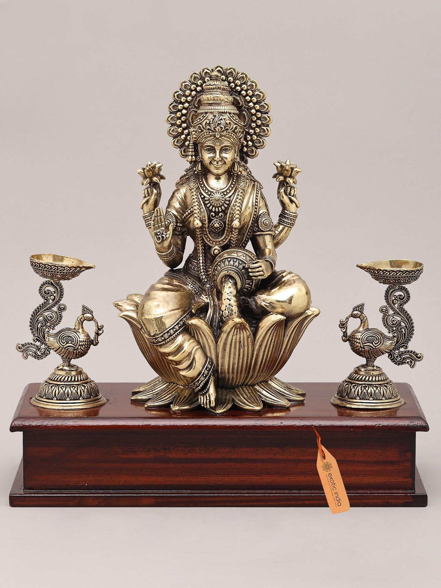 9" Superfine Four Armed Goddess Lakshmi with Peacock Lamp and Wood Base | Brass Idol: The Embodiment of Prosperity and Auspiciousness This exquisite brass idol captures the divine essence of Goddess Lakshmi, the revered Hindu deity of wealth, fortune, prosperity, and auspiciousness. Se
