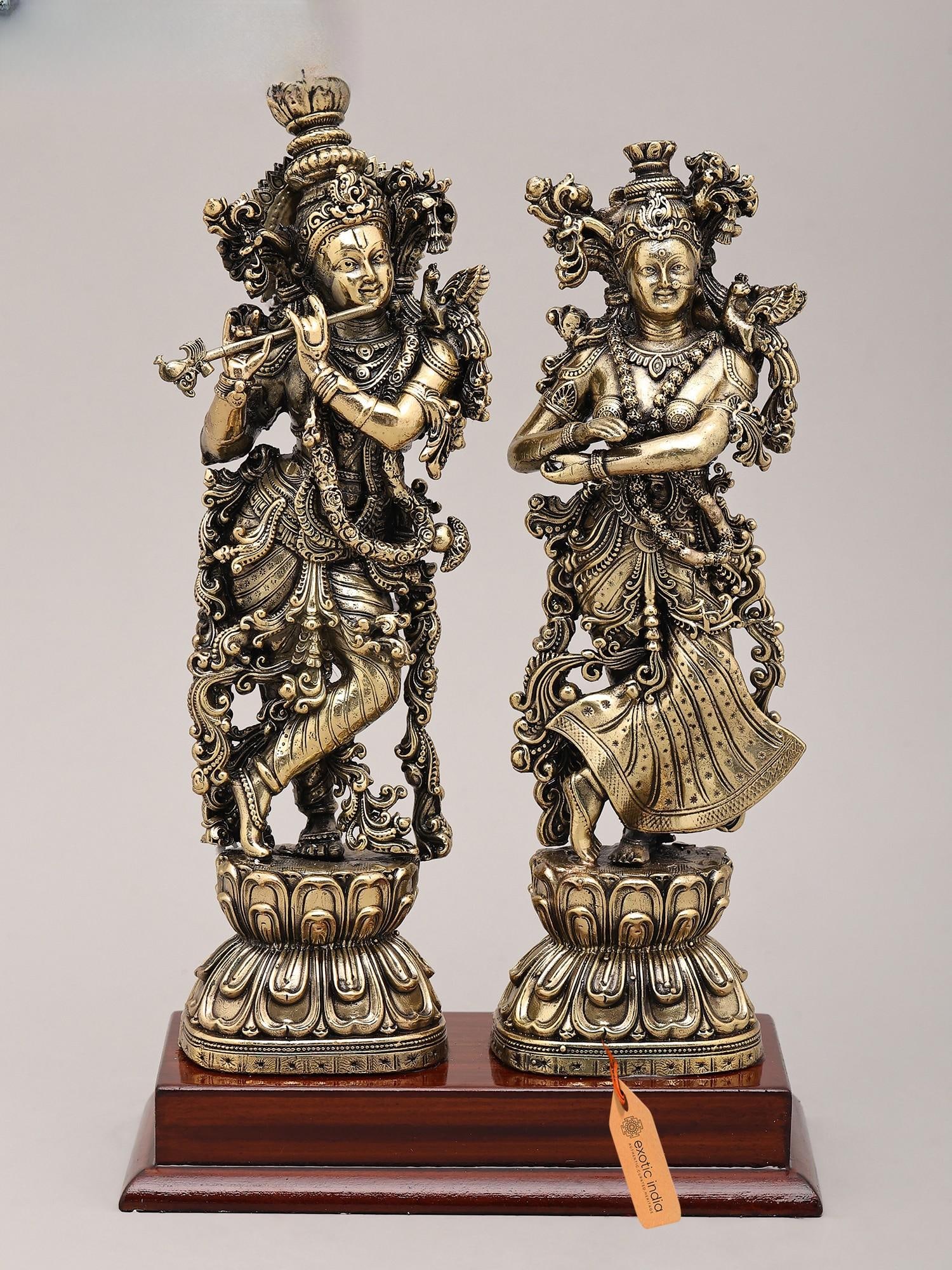Superfine Divine Pair: Lord Krishna and Radha Standing on Lotus with Wood Base | Brass Idol: The Divine Embrace: A Testament to Eternal Love This exquisite brass idol captures the quintessential moment of divine love between Lord Krishna and Radha, two figures revered for their