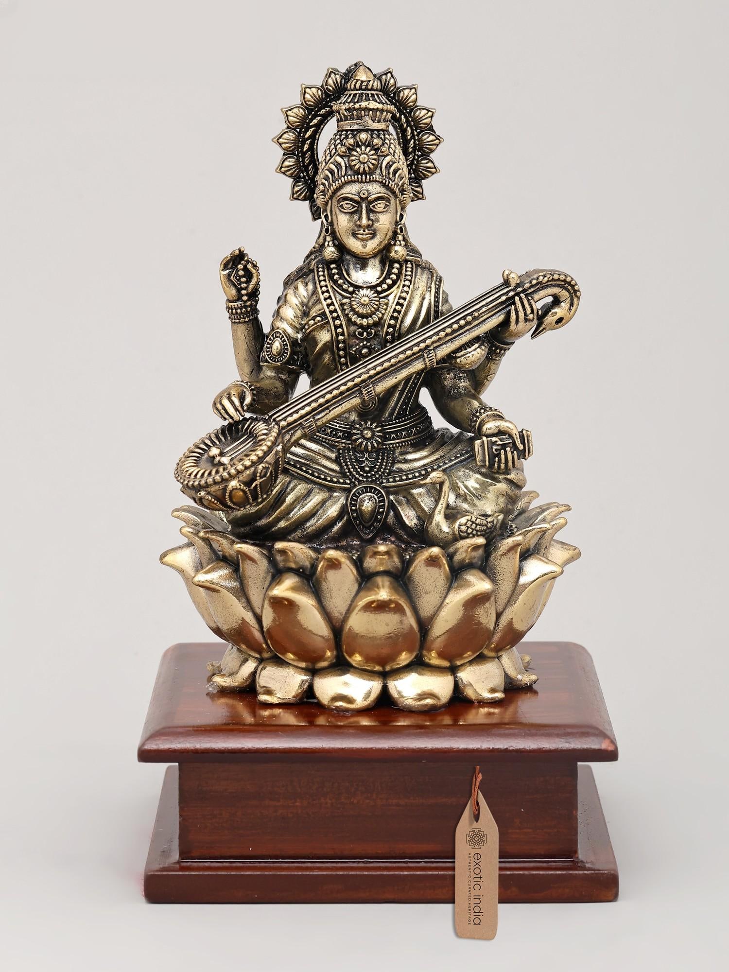 7" Superfine Chaturbhuja Goddess Saraswati Seated on Lotus with Wood Base | Brass Sculpture: A Divine Embodiment of Knowledge and Arts This exquisite brass sculpture captures the essence of Goddess Saraswati, the Hindu deity of knowledge, music, arts, wisdom, and nature. Revered as the