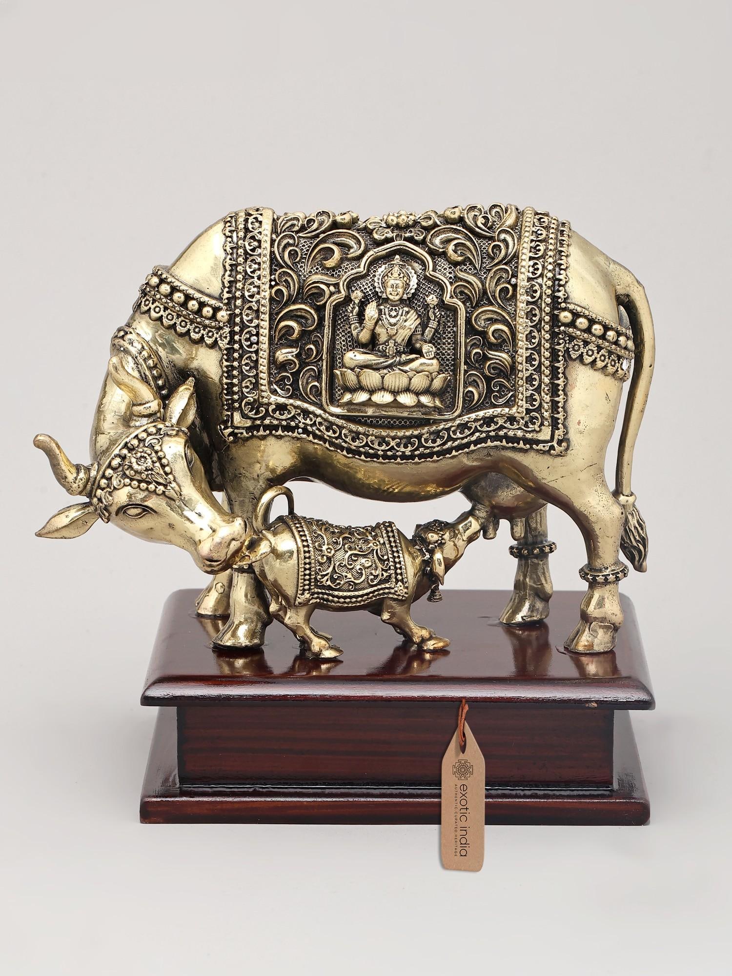 6" Superfine Goddess Kamdhenu Cow and Calf with Goddess Lakshmi Carving on Wood Base | Brass Statue: The Nurturing Divine: Kamadhenu and Calf This exquisite brass statue captures the essence of abundance, nurture, and divine grace. It depicts Kamadhenu, the celestial wish-fulfilling cow, with her cal