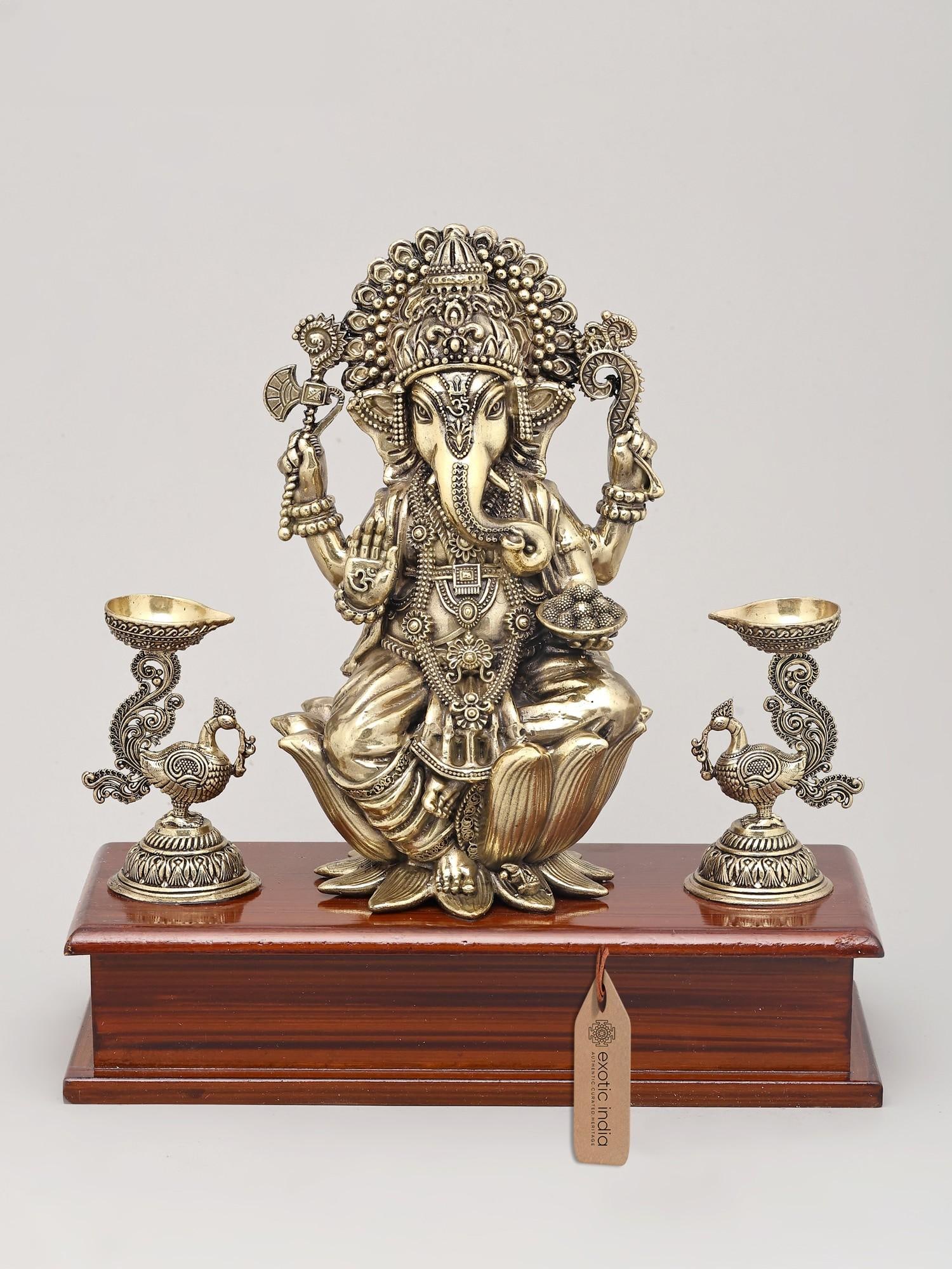 9" Superfine Lord Ganesha Seated on Lotus with Oil Lamps and Wood Base | Brass Sculpture: The Auspicious Presence of Ganapati This exquisite brass sculpture captures the divine essence of Lord Ganesha, the remover of obstacles and the bestower of prosperity. Seated serenely on a blooming l