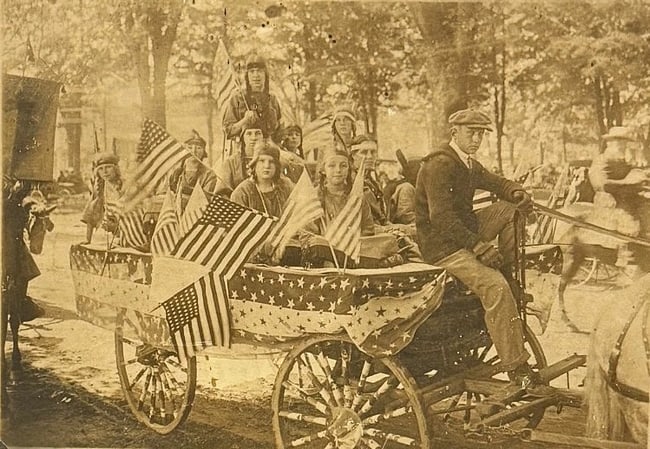 Patriotic Photograph Women Dressed as Native Americans w American Flags 1912: Patriotic albumen photograph of a parade float with women and girls dressed as Native Americans holding American Flags. Photo is in good condition with some staining and a corner tear. Board attached 