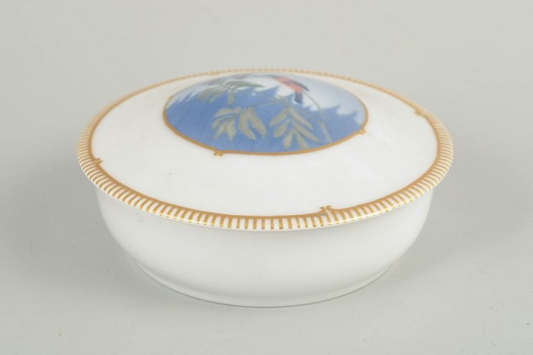 Royal Copenhagen, Denmark. Art Deco porcelain lidded dish with gilt rim and bird motif. Approx.: Royal Copenhagen, lidded dish. Art Deco lidded dish in white-glazed porcelain with a wide gilt rim and a central hand-painted bird motif in blue and green tones. First quality. Model 1340/2. Origin: D