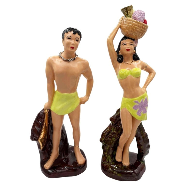 Mid-Century Hand Painted Hawaiian Hula Girl and Male Surfer Ceramic Statue Set: This pair of vintage mid-century ceramic figurines depicts a stylized Polynesian man and woman. Hand-painted with a glossy finish, the man holds a paddle while the woman balances a basket on her head,