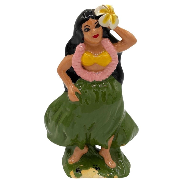 Mid-century "Hawaiian" Hula Girl in Grass Skirt Ceramic Statue: 1955 Mid-century Era pink and yellow hula girl ceramic statue. The statue features a beautifully detailed hula girl holding a flower in her hair with the word "Hawaiian" along the base. It comes in a 